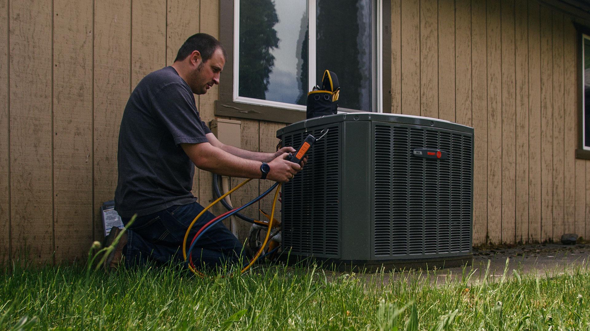 A&E HVAC tech kneeling in grass, working on outdoor unit