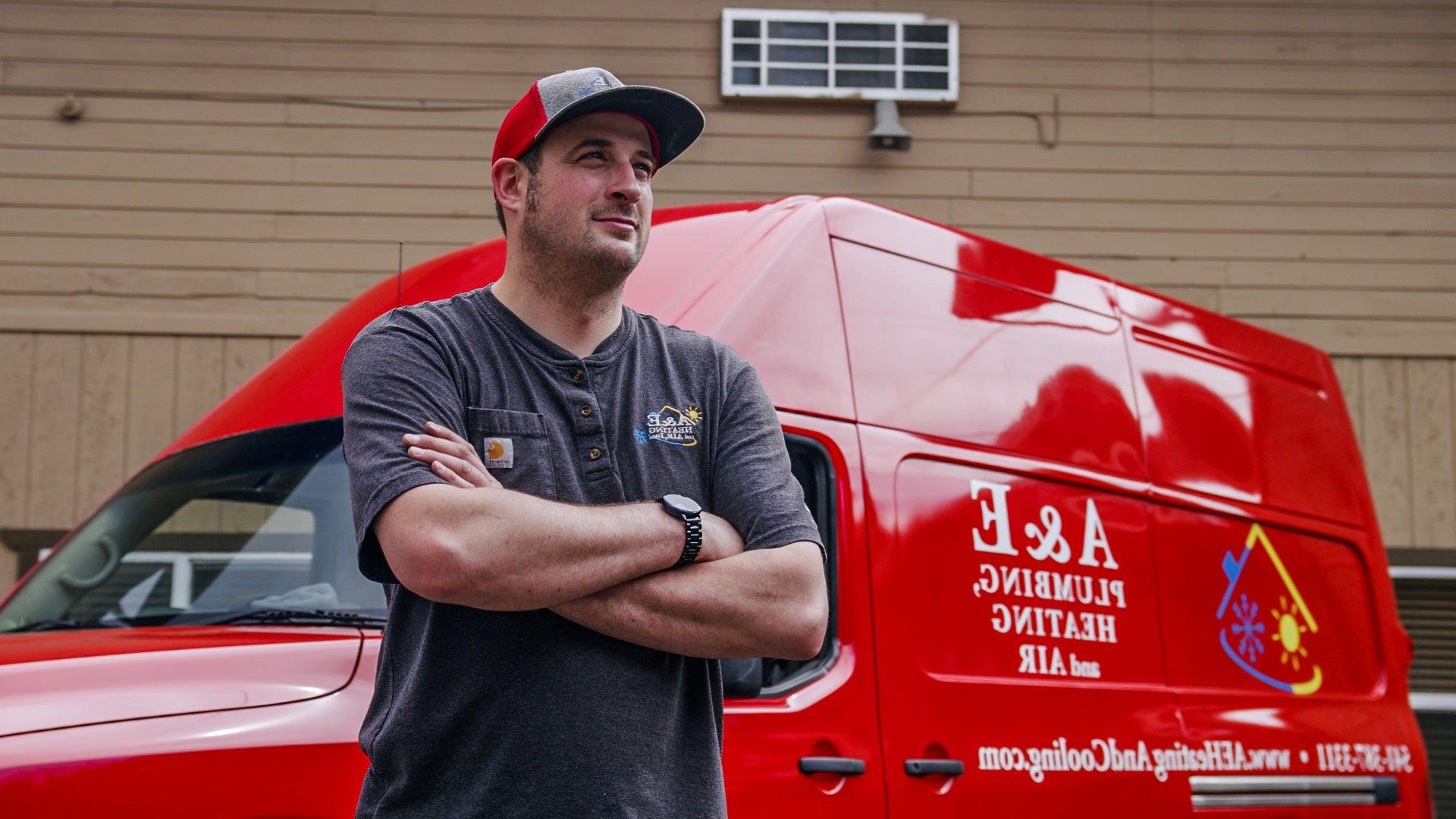 A&E tech standing in front of a bright red A&E company van, his arms folded and a small smile on his face.